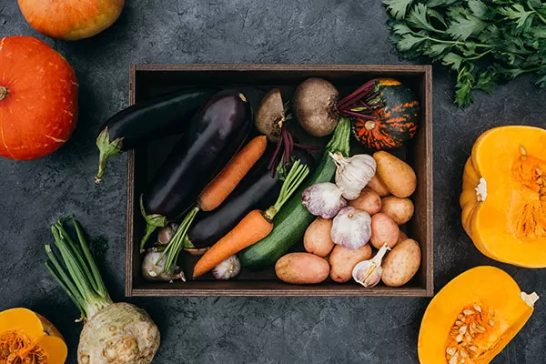 A crate of vegetables on a black background surrounded by pumpkins and greenery